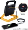 A portable LED work light is shown charging a smartphone, demonstrating its power bank functionality. Produktbild-Vorschau