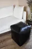 A black leather pouf sits on a wooden floor next to a white sofa. Produktbild-Vorschau