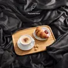 A cozy breakfast scene with a croissant, coffee, and cookies laid out on a wooden tray, all nestled within a soft, dark gray blanket. Produktbild-Vorschau