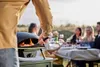 People enjoying a meal outdoors with a pizza oven in the foreground. Produktbild-Vorschau