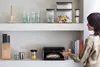 A woman is reaching into a black bread bin on a shelf, which contains a loaf of bread. The shelf also holds other kitchen items like glasses, jars, and a knife block. Podgląd zdjęcia produktu