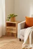 A wooden nightstand with a woven drawer front sits next to a chair with an orange pillow and a cream blanket. Produktbild-Vorschau