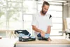 A man is working on a workbench with a sander and a box of accessories. Voir les images du produit