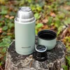 A light green thermos bottle and its parts are placed on a tree stump in an outdoor, natural setting with blurred foliage in the background. Voir les images du produit