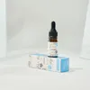 A small glass bottle with a dropper cap sits on top of its blue and white cardboard packaging, against a plain white background with subtle textured elements on the sides. Náhľad obrázku produktu