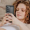 A woman is lying down and holding a smartphone with a black case. Produktbild-Vorschau
