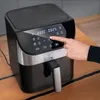 A hand is shown pressing buttons on the digital display of a black and silver air fryer, which is placed on a wooden table. Podgląd zdjęcia produktu