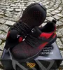 A pair of black and red safety shoes are placed on a box, which is on a cobblestone surface. Produktbild-Vorschau
