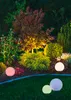 A garden at night, illuminated by various lights, including a tree lit from below and several spherical outdoor lights. Produktbild-Vorschau
