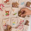 A hand reaches into a fabric advent calendar to pull out a small brown paper bag. The calendar is decorated with various Christmas-themed illustrations. Produktbild-Vorschau