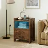 A wooden nightstand with drawers and an open shelf, featuring a vase with flowers, a phone charging station, and a radio. It is placed next to a sofa in a room with a floor lamp. Produktbild-Vorschau