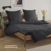 A bed with dark gray bedding, pillows, and a wooden frame, with a plant and a vase with dried flowers in the background. Produktbild-Vorschau
