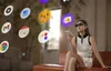 A woman wearing AR glasses sits on a couch, holding a smartphone, with holographic app icons floating around her. Anteprima dell’immagine prodotto
