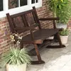 A wooden rocking bench sits on a patio next to a brick wall, with potted plants around it. Náhľad obrázku produktu