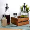 A set of oil and vinegar bottles and shakers arranged on a light blue cloth on a wooden surface, with a potted plant in the background. Produktbild-Vorschau