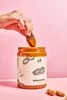 A hand is dipping a date into a jar of peanut butter. Produktbild-Vorschau
