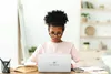 A young girl wearing glasses is focused on a laptop in front of her, sitting at a desk with books and pens. Produktbild-Vorschau