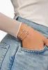 A close-up of a person's wrist wearing two delicate silver bracelets, one with an infinity symbol and the other with a heart charm, against a backdrop of denim jeans. Anteprima dell’immagine prodotto