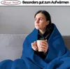 A woman is sitting on a couch wrapped in a blue blanket, holding a mug. Produktbild-Vorschau