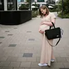 A pregnant woman stands on a paved walkway, holding a black diaper bag. Produktbild-Vorschau