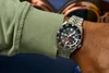 A person's wrist wearing a watch, with a green sleeve visible. Produktbild-Vorschau