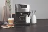 A coffee machine is shown with a glass of coffee and a jug of milk, along with coffee beans scattered on the table. Produktbild-Vorschau