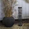 A black solar garden light with a decorative pattern stands next to a large black planter filled with dried grass, set against a white wooden wall. Produktbild-Vorschau