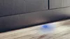 A close-up shot of the bottom of a dark gray appliance, showing a small, glowing blue light on the wooden floor beneath it. Produktbild-Vorschau