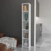 A tall, narrow white bookshelf filled with books and decorative vases, placed next to a brown leather armchair with a patterned throw blanket. In the background, a modern kitchen area is visible. Produktbild-Vorschau