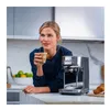 A woman is holding a cup of coffee next to an espresso machine. Produktbild-Vorschau