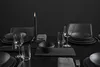 A dark, elegantly set dining table with black plates, glassware, and a lit black candle. Produktbild-Vorschau