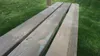 A close-up view of wooden planks of a bench, with grass in the background. Produktbild-Vorschau