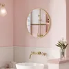 A modern bathroom with pink walls, a white sink, and gold fixtures, featuring a round gold-framed mirror reflecting a shower. Produktbild-Vorschau