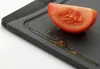 A close-up of a slice of tomato on a dark gray cutting board with some liquid. The cutting board has a raised edge to catch liquids. Produktbild-Vorschau