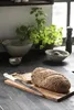 A loaf of bread and a knife on a wooden cutting board, with kitchen items and a plant in the background. Produktbild-Vorschau