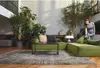 A woman is vacuuming a living room with a large green sectional sofa and many plants. Náhled obrázku produktu