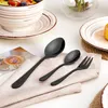 A set of black cutlery is arranged on a light-colored napkin, with a bowl of salad and plates in the background. Produktbild-Vorschau