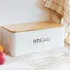 A white bread box with a wooden lid sits on a wooden surface, with a checkered cloth draped in the background and two apples in the foreground. Podgląd zdjęcia produktu