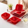 A person is arranging a set of red square plates and bowls on a white table, with a small potted plant and a salt and pepper grinder in the background. Náhled obrázku produktu