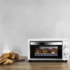 A white mini oven with croissants inside is on a wooden table, with more croissants and cookies next to it. Produktbild-Vorschau