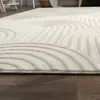 A close-up view of a textured white rug with a geometric pattern, lying on a dark wooden floor. The background shows a blurred interior scene with furniture. Anteprima dell’immagine prodotto