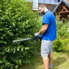 A man is trimming a hedge with a cordless hedge trimmer in a garden. There is a house in the background. Náhled obrázku produktu