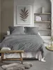 A bedroom scene with a neatly made bed, featuring grey bedding and pillows. A wooden stool with books sits in front of the bed, and a fluffy rug is on the floor. Produktbild-Vorschau