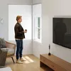 A young man stands in a modern living room, holding a cup and looking out a window. A TV is mounted on the wall, and a wooden media console is below it. A wicker chair with a blanket is to the left. Produktbild-Vorschau
