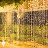 A curtain of warm white LED string lights hanging outdoors, possibly on a balcony or patio, with greenery in the background and a USB connector visible in the foreground. Produktbild-Vorschau