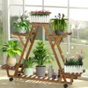 A wooden plant stand filled with various potted plants is placed in front of a large window, with a blurred outdoor scene visible through the glass. Produktbild-Vorschau
