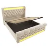 A beige upholstered bed frame with the mattress support raised, revealing storage space underneath. Yellow LED lighting is integrated into the headboard and base. Produktbild-Vorschau