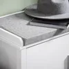 A close-up view of a white shoe bench with a gray upholstered seat, featuring a gray hat and a book placed on top. Náhľad obrázku produktu