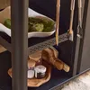 A close-up view of a black outdoor serving cart with various items on its shelves, including artichokes in a white bowl, kitchen utensils, and jars of spices on a wooden tray. Produktbild-Vorschau