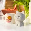 A cute gray cat figurine sits next to a mug with a cookie graphic, on a table with strawberries and tulips in the background. Voir les images du produit